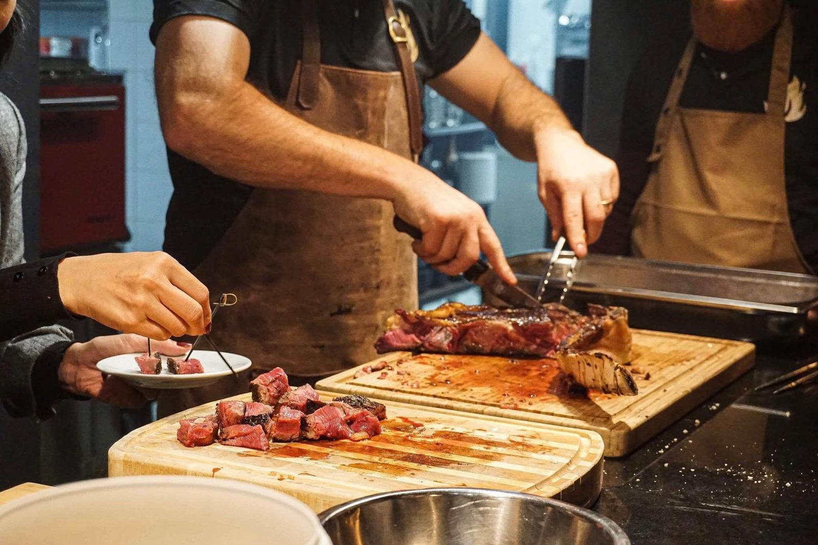 Trois personnes portant des tabliers préparent et servent des tranches de steak sur des planches à découper en bois. L'une d'elles découpe la viande, une autre sert les morceaux et une troisième se tient à proximité. La scène se déroule dans une cuisine.
